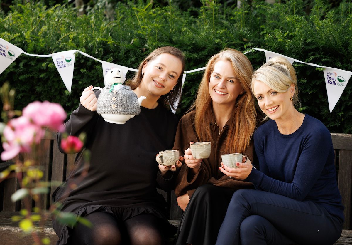 Three women sitting on a bench, smiling, holding mugs and a teapot with a cozy. Bunting hangs behind them in a garden setting.